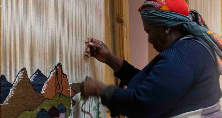 Een bejaarde vrouw die een kleurrijk textieltuk weeft met een natuurtafereel.