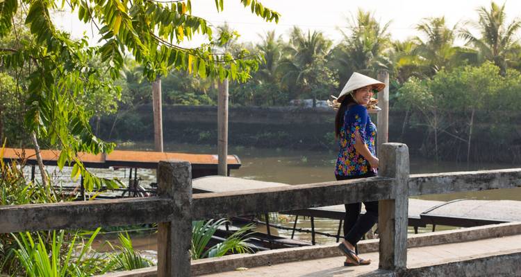 Una mujer con una camisa floral y un sombrero cónico está de pie en un puente sobre el río rodeada de palmeras.