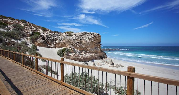 Una playa pintoresca con paseo marítimo de madera, acantilados rocosos y océano azul.