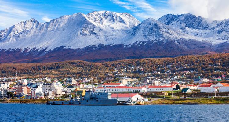 Naval vessel and town buildings along the shoreline with towering snow-dusted peaks behind