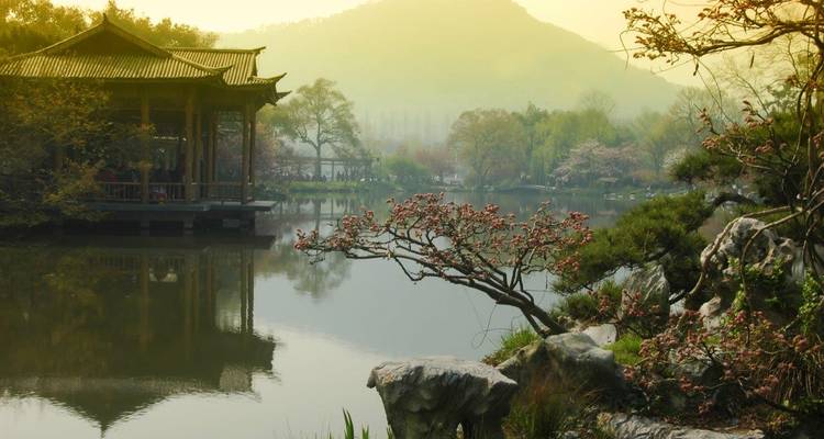 Scène paisible au bord du lac avec un pavillon traditionnel et des montagnes brumeuses en arrière-plan.