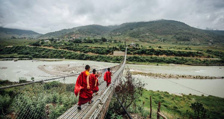 Des moines en robes rouges traversant un pont suspendu au-dessus d'une rivière.