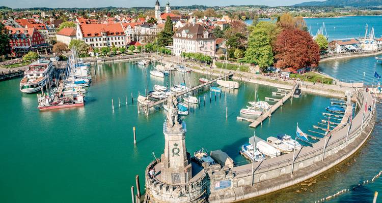 Drohnenaufnahme des Lindauer Hafens mit Leuchtturm, Löwenstatue und türkisfarbenem Wasser, umgeben von bunten Dächern der Stadt.