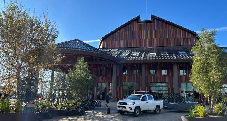 Centro de ciclismo contemporáneo revestido de madera con bicicletas estacionadas, camioneta y visitantes bajo un cielo azul despejado.