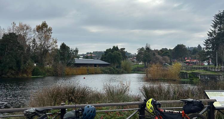 Vista nublada a través de un estanque tranquilo bordeado por árboles otoñales con cascos de ciclismo y equipo descansando sobre una barandilla de madera.