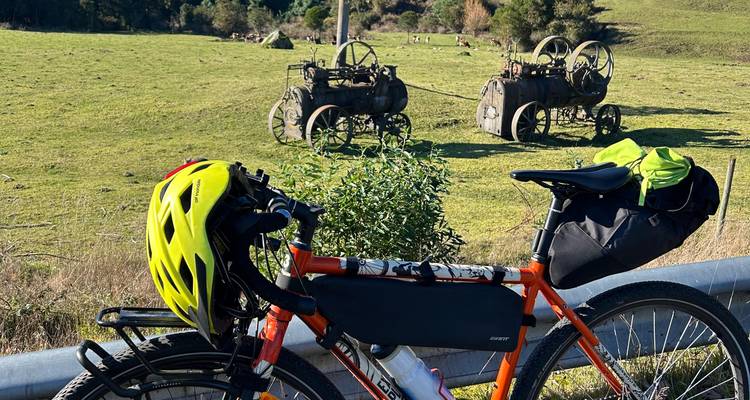 Bicicleta de turismo naranja con casco y alforjas estacionada junto a un pasto rural que contiene dos tractores de vapor antiguos.