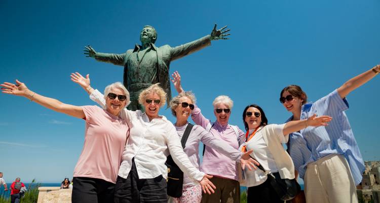 Un groupe joyeux de femmes pose les bras écartés à côté de la statue en bronze du chanteur Domenico Modugno sous un ciel bleu éclatant.