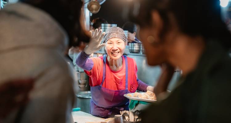 Alegre vendedor de mercado usando guantes saluda mientras sirve comida dentro de un puesto bullicioso