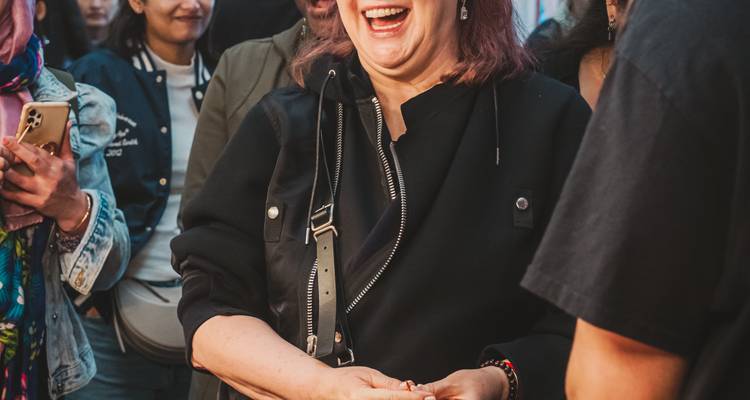 Mujer riendo en chaqueta negra disfrutando un recorrido por el mercado en interiores con multitud borrosa a su alrededor