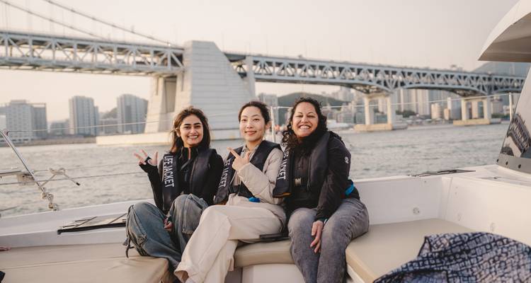 Tres mujeres con chalecos salvavidas se relajan en un yate con el puente Gwangan de Busan y el horizonte de la ciudad detrás de ellas