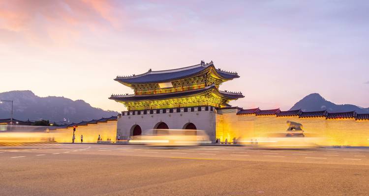 Puerta de Gwanghwamun iluminada de Seúl al atardecer con tráfico difuminado por el movimiento en primer plano y montañas silueteadas detrás