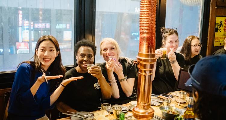 Amigos disfrutando barbacoa coreana y bebidas dentro de un restaurante, sonriendo a la cámara
