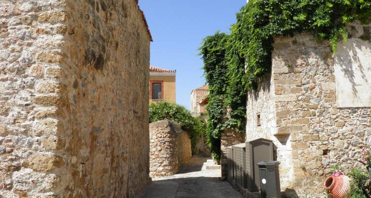 Ruhige, sonnenbeschienene Steinstraße zwischen historischen Gebäuden, die mit Efeu überwachsen sind, in einem griechischen Dorf