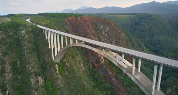 Drohnenaufnahme der langen Bloukrans-Bogenbrücke, die sich über eine tiefe grüne Schlucht mit Bergen im Hintergrund erstreckt.