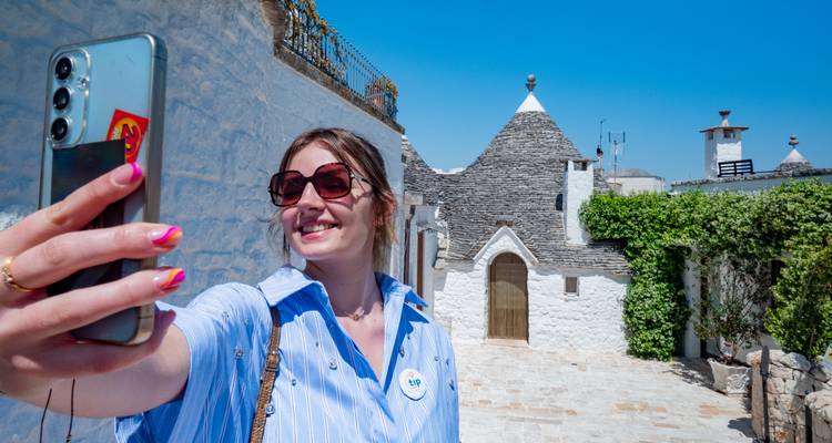 Un voyageur souriant prend un selfie parmi les maisons trulli blanchies à la chaux sous un ciel bleu éclatant.