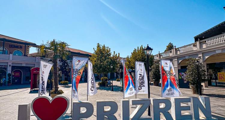 Plaza de la ciudad con banderas y letrero de 'Amo Prizren' en un día soleado.