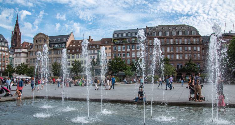 Fuentes en una plaza con edificios históricos europeos y torre puntiaguda.