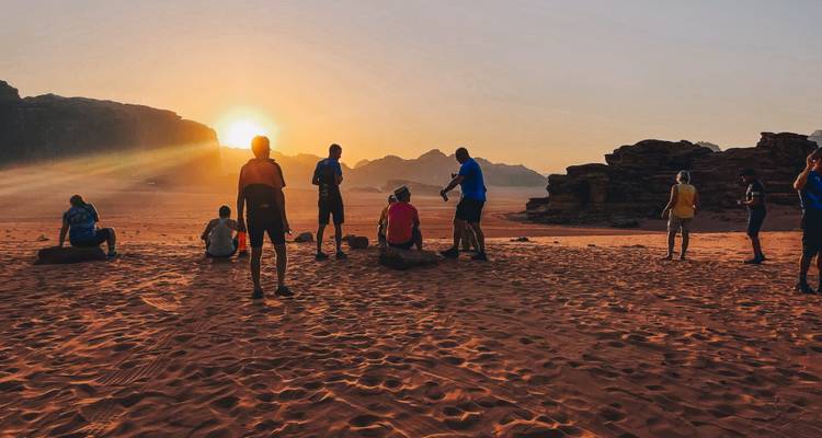 Travellers gather on orange sand to watch sunrise over rugged Wadi Rum landscape.