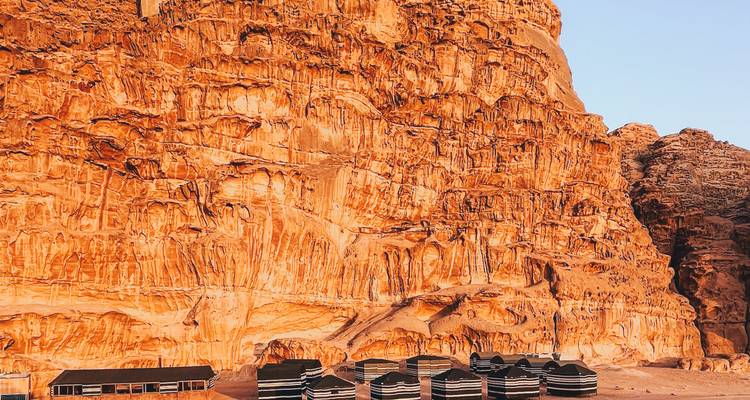 Row of traditional black-striped desert tents dwarfed by dramatic red sandstone cliff.