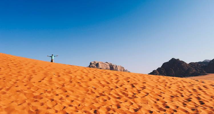 Traveller raises arms triumphantly atop a vast orange sand dune beneath deep blue sky.