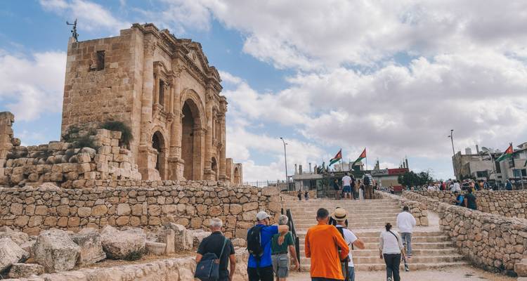 Crowds explore the ancient stone arch and ruins of Jerash under partly cloudy sky.