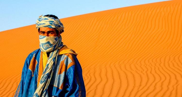 Ein Mann in traditioneller marokkanischer Kleidung, der vor einer Sanddüne in der Wüste steht.