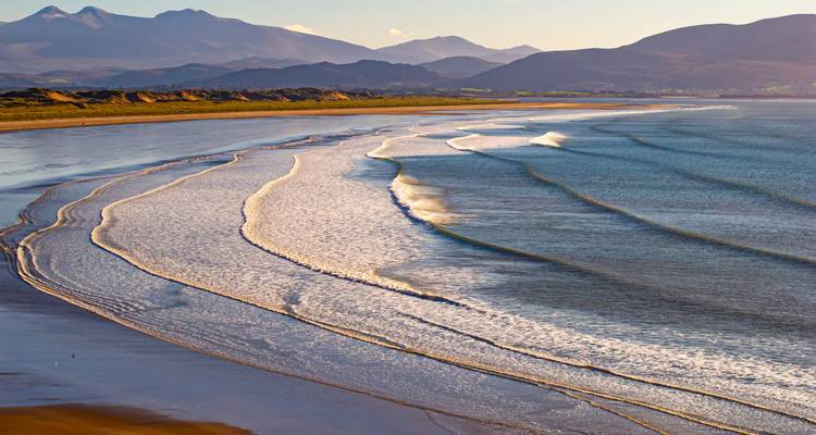 Wellen, die sanft an einen Sandstrand mit Bergblick spülen.