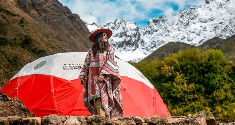 Mujer en atuendo tradicional de pie junto a una tienda con picos nevados