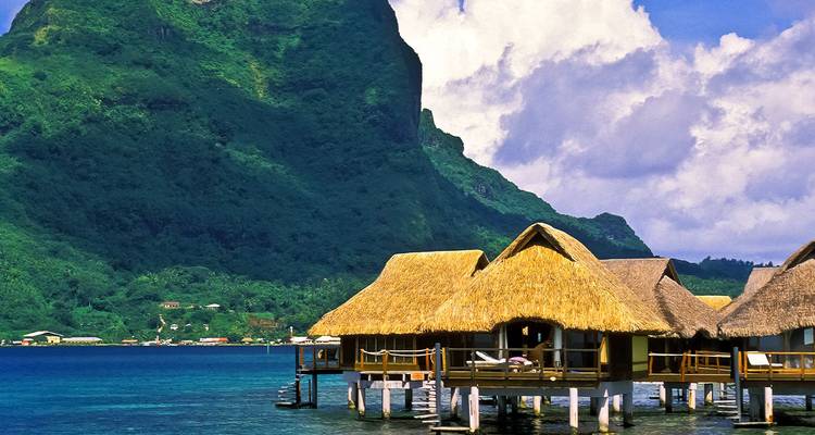 Strohdächer über dem Wasser errichtete Bungalows liegen über einer türkisfarbenen Lagune mit einer üppigen grünen Bergkulisse im Hintergrund.