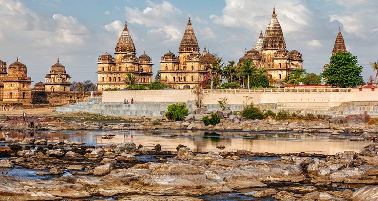 Serie de cenotafios ornamentados de Orchha reflejados en el río Betwa con rocas en primer plano.