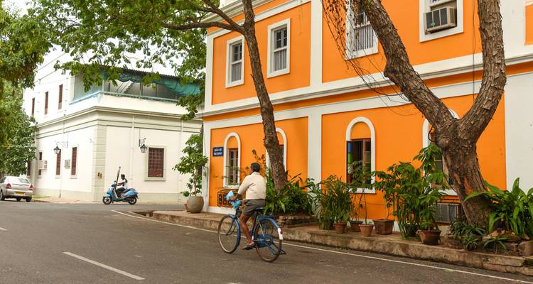 Una persona en bicicleta pasando por un vibrante edificio colonial de color naranja con árboles bordeando la calle.