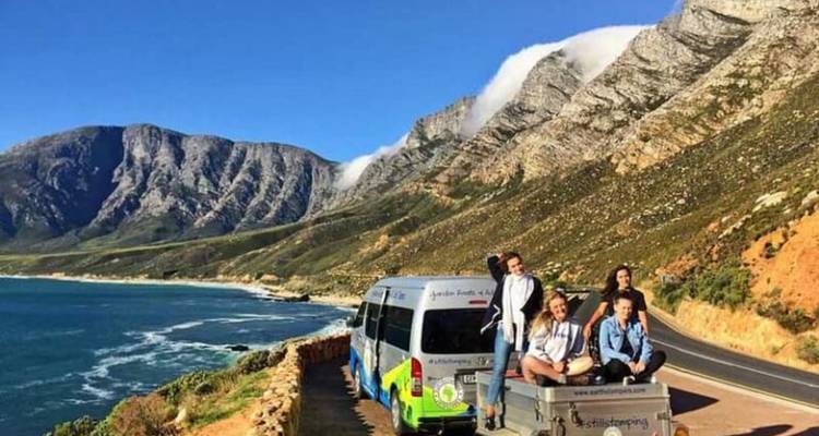 Junge Reisende sitzen auf einem Anhänger neben einem bunten Kleinbus auf einer Klippenstraße mit Blick auf dramatische Küstenberge und das Meer
