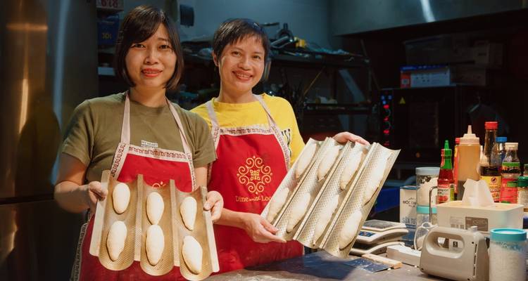Two people holding trays with raw baguettes in a kitchen.