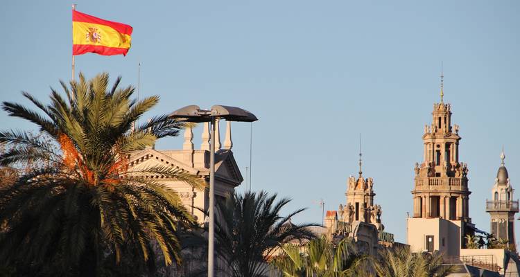 Bandera española ondeando sobre edificios ornamentados en una plaza de la ciudad.