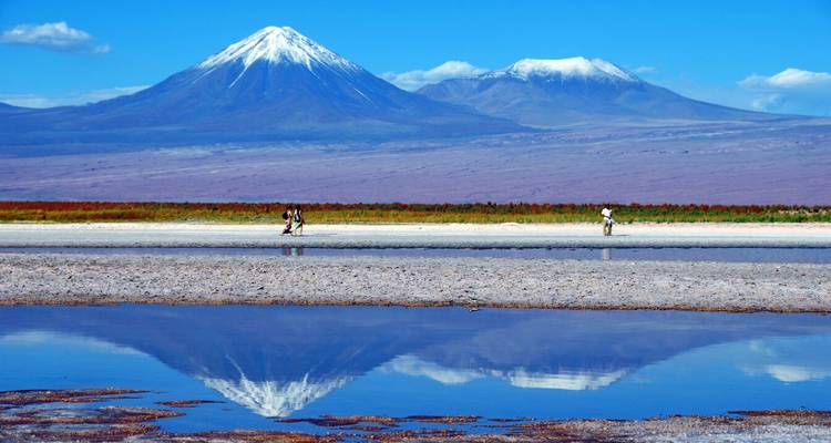 Bergen weerspiegeld in water met mensen die langs de rand lopen.
