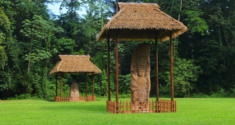 Alte Steinmetzarbeiten in einer offenen Grasfläche, umgeben von dichtem Dschungel.