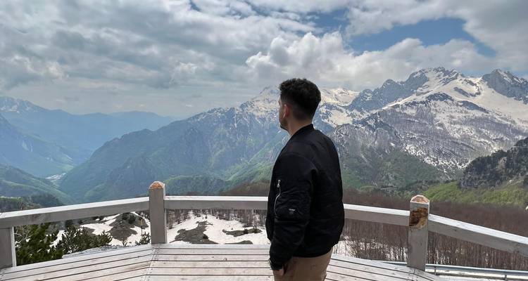 Une personne debout sur une plateforme en bois regardant un paysage de montagne avec des sommets enneigés.
