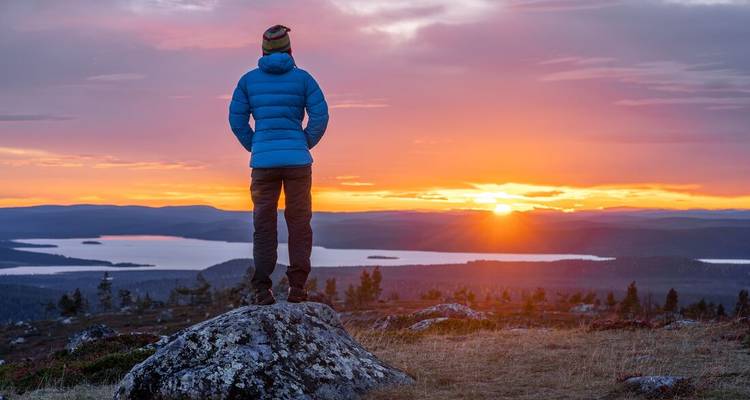 Eine Person, die auf einem Felsen steht und einen Sonnenuntergang über einem See betrachtet.