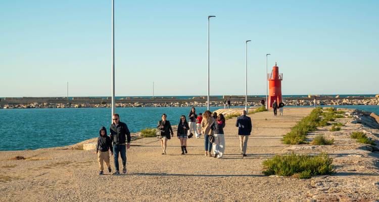 Les familles et les couples se promènent le long d'une jetée en pierre vers un petit phare rouge au bord de la mer.