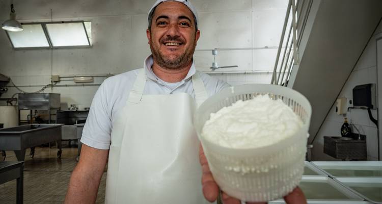 Un fromager souriant présente un panier de ricotta fraîche dans une salle de production lumineuse.