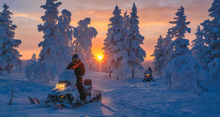 Deux personnes faisant de la motoneige à travers une forêt enneigée et ensoleillée.