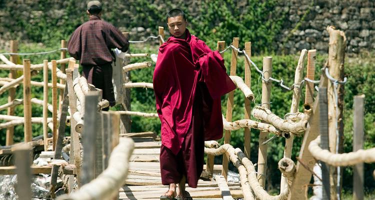 Mönch in roter Robe, der über eine Holzbrücke geht, mit einer anderen Person im Hintergrund.