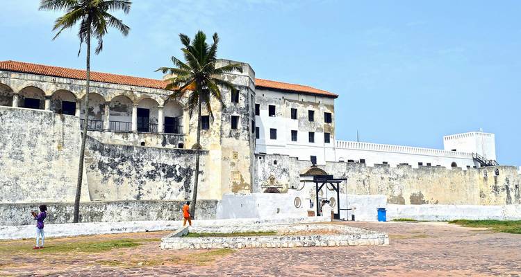 Les murs blancs usés du château historique d'Elmina et ses arcades voûtées avec palmier et enfants jouant dans la cour avant.