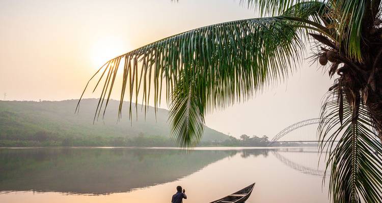 Peaceful river scene at sunrise with a lone man in a canoe, a sweeping palm frond, misty hills and an arched steel bridge in the distance.