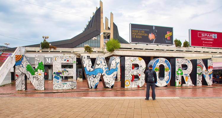 Un grand panneau coloré 'NOUVEAU-NÉ' avec des motifs détaillés et une personne debout devant, entouré de bâtiments.