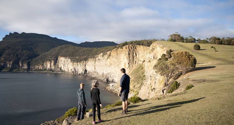 Drie mensen die op een grasrijke heuvel staan en uitkijken over een schilderachtige kustlijn met kliffen en oceaan.