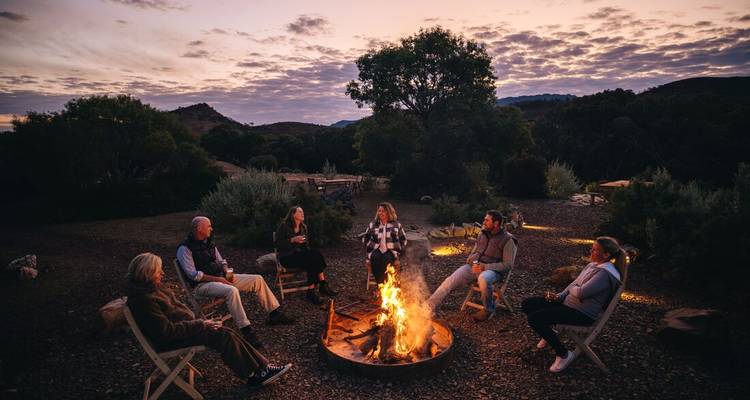 Mensen die rond een kampvuur zitten met bergen op de achtergrond.