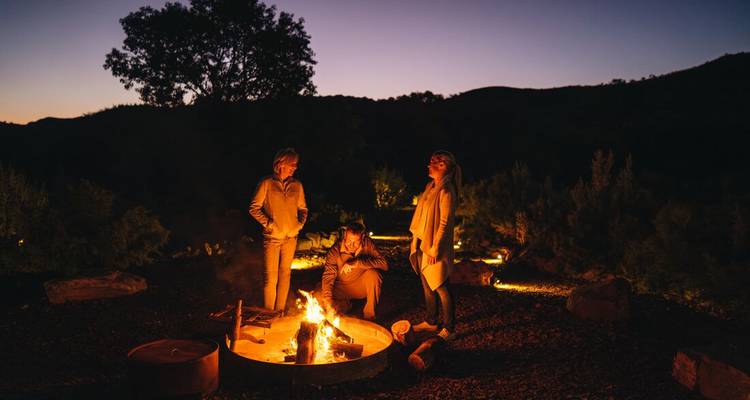 Drie mensen genieten van een kampvuur in de avond in een bergachtig landschap.