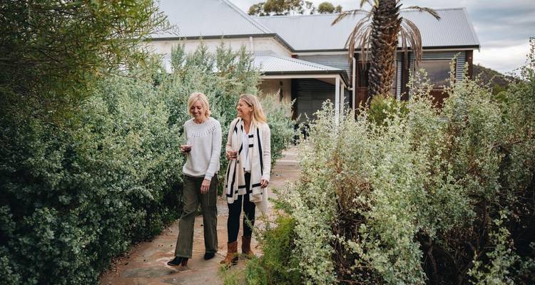 Twee vrouwen lopen door een tuin bij een landelijk huis, houden drankjes vast en glimlachen.