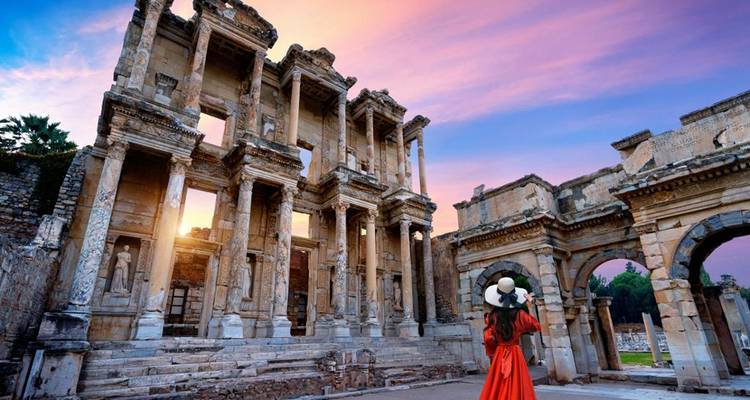 Mujer con vestido rojo de pie frente a ruinas antiguas al atardecer.